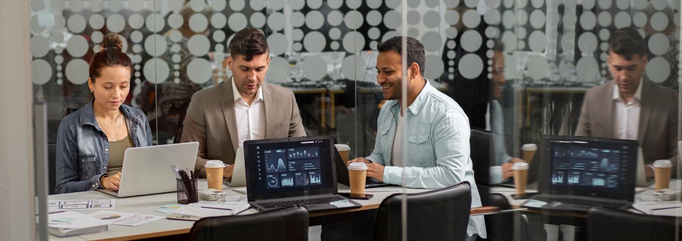 Kobieta i dwóch mężczyzn pracujący na komputerach przy wspólnym stole w przestrzeni biurowej