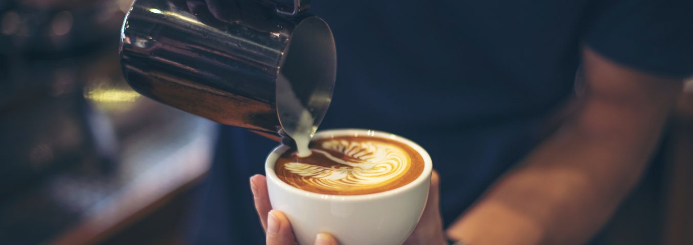 Zbliżenie na dłonie baristy, który nalewa do filiżanki mleko tworząc wzory na kawie tzw. latte art.