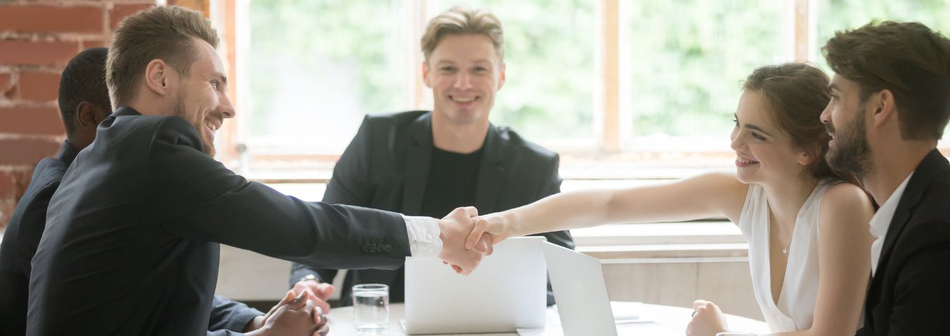 Grupa ludzi siedząca przy stole. Kobieta i mężczyzna podają sobie ręce