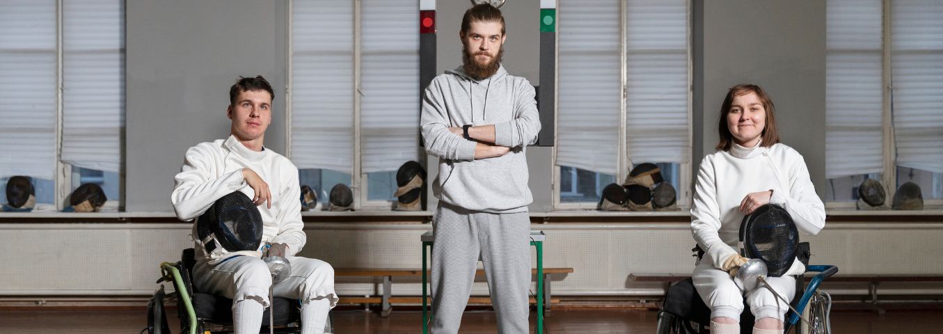 Kobieta i mężczyzna na wózkach inwalidzkich przygotowani do treningu szermierki. Między zawodnikami stoi trener.